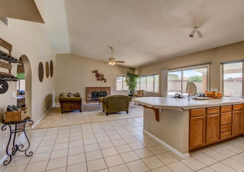 Spacious open-plan living area with beige tiled floor and light beige walls. The room features a kitchen island with wooden cabinets and a tiled countertop, decorated with a fruit basket and tableware. Adjacent to the kitchen is a living room area with two green upholstered armchairs, a sofa, a ceiling fan with light, a brick fireplace, and wall decorations. Large windows and sliding glass doors provide natural light and a view of the outdoor area.