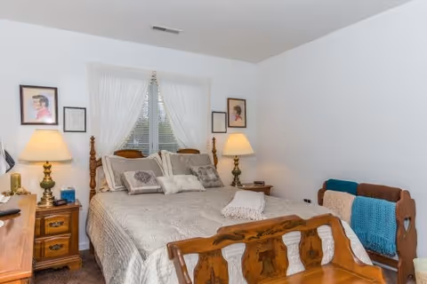 A cozy bedroom with a wooden bed frame and matching nightstands on either side. The bed is made with a beige quilt and several decorative pillows. Two table lamps with beige shades sit on the nightstands. A window with white curtains is centered behind the bed. There are framed pictures on the walls beside the window. A wooden bench with a blue and beige knitted blanket is placed at the foot of the bed.