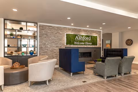 A modern and cozy living room area in The Ashford at Sturbridge featuring a stone accent wall with a green sign that reads 'The Ashford Welcome Home'. The room has comfortable seating including beige armchairs, blue and gray chairs, a round ottoman, and a built-in fireplace. There is also a shelving unit with decorative items and plants.