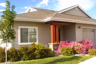 Exterior view of a single-story residential building with beige siding, a brown shingled roof, a garage door, a window, and a small porch area with red pillars. There are green bushes and pink flowering plants in front of the building under a blue sky with some clouds.