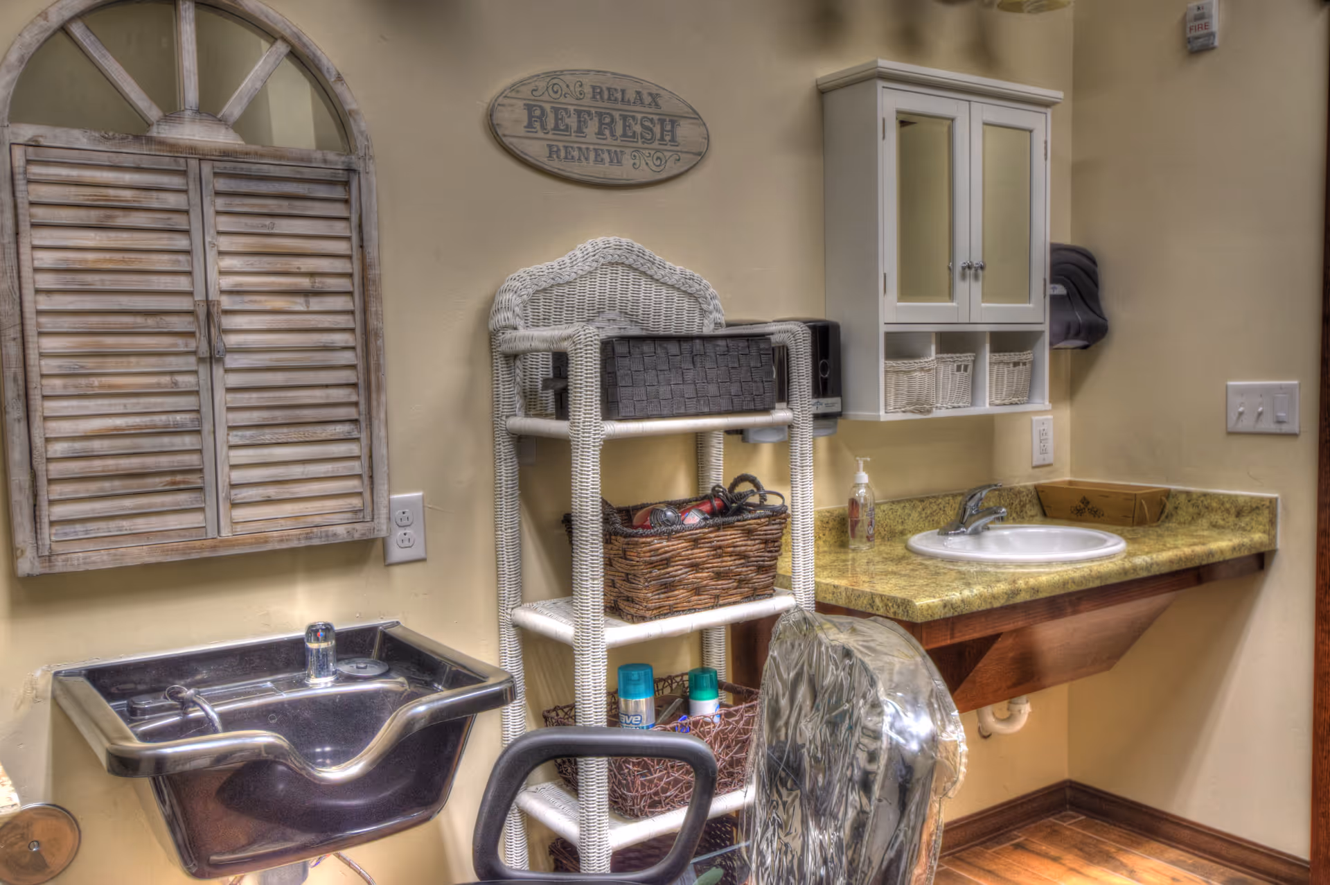 Interior of a small salon or grooming area with a black shampoo sink, a white wicker shelving unit holding baskets and hair products, a countertop with a sink and soap dispenser, a white wall cabinet with mirrored doors, and a decorative wooden wall piece shaped like a window with shutters. A sign above the shelving unit reads 'Relax Refresh Renew'.