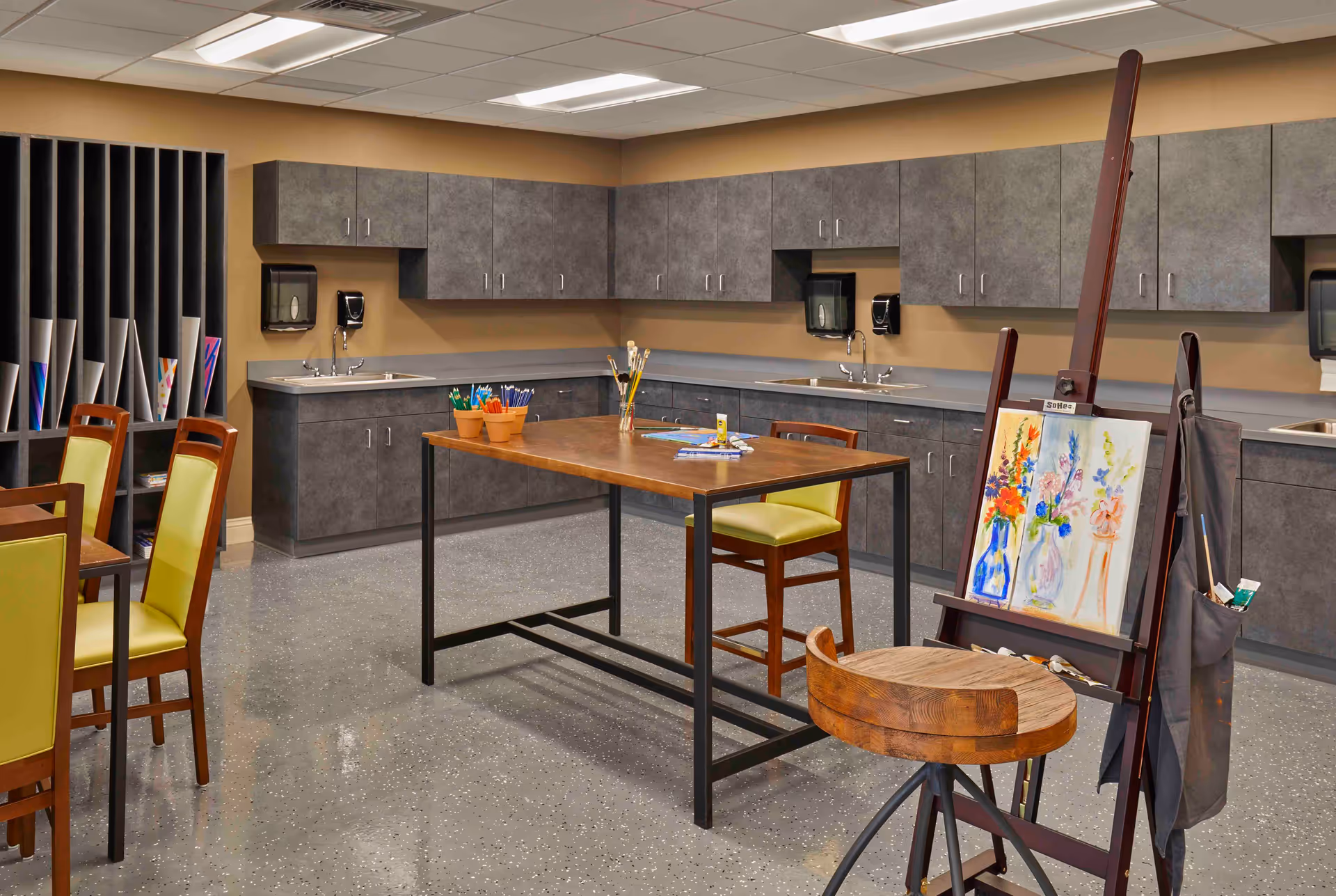 An art room with a wooden table and chairs, art supplies including paintbrushes and colored pencils on the table, an easel holding a painting of colorful flowers, and gray cabinets with sinks along the walls.