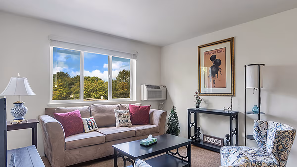 Well-lit living room with a beige sofa, colorful throw pillows, a coffee table, patterned armchair, and a large window showing trees.
