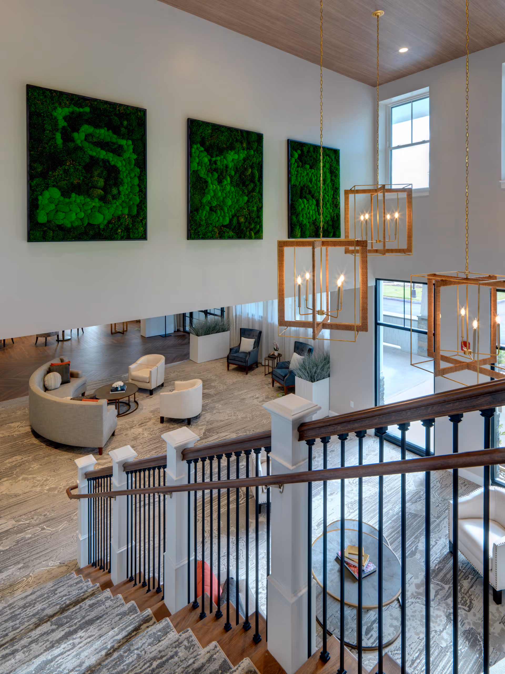 View from the top of a staircase overlooking a spacious, modern living room area with a curved sofa, armchairs, and coffee tables. The room features large windows, three green moss wall art pieces, and three pendant light fixtures with a geometric design hanging from a wooden ceiling.
