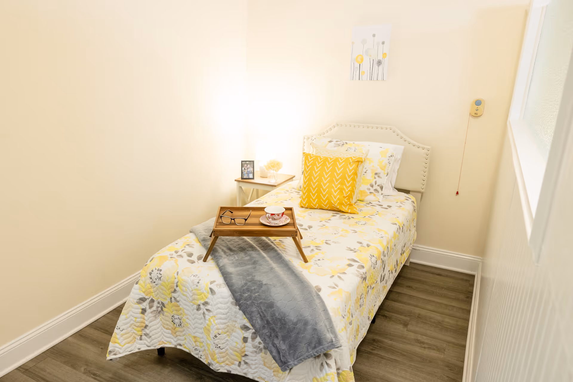 A small, cozy bedroom with a single bed covered in a floral yellow and gray bedspread, a yellow decorative pillow, and a gray throw blanket. A wooden tray with a pair of glasses and a teacup sits on the bed. There is a small side table with a lamp, a framed photo, and a decorative item. The walls are light-colored, and a small piece of floral artwork hangs above the bed. A pull cord is visible on the wall near the bed.