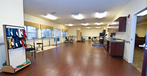 A spacious rehabilitation or physical therapy room with wooden flooring, exercise equipment including stationary bikes and parallel bars, a rack with various exercise bands and weights, large windows letting in natural light, and cabinets with a countertop along one wall.