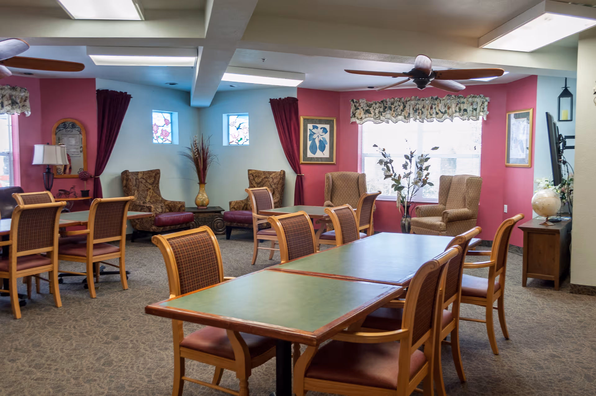 A cozy assisted living common area with multiple tables and chairs arranged for group seating. The room features comfortable armchairs near windows with floral curtains and decorative wall art. Ceiling fans and soft lighting create a welcoming atmosphere.