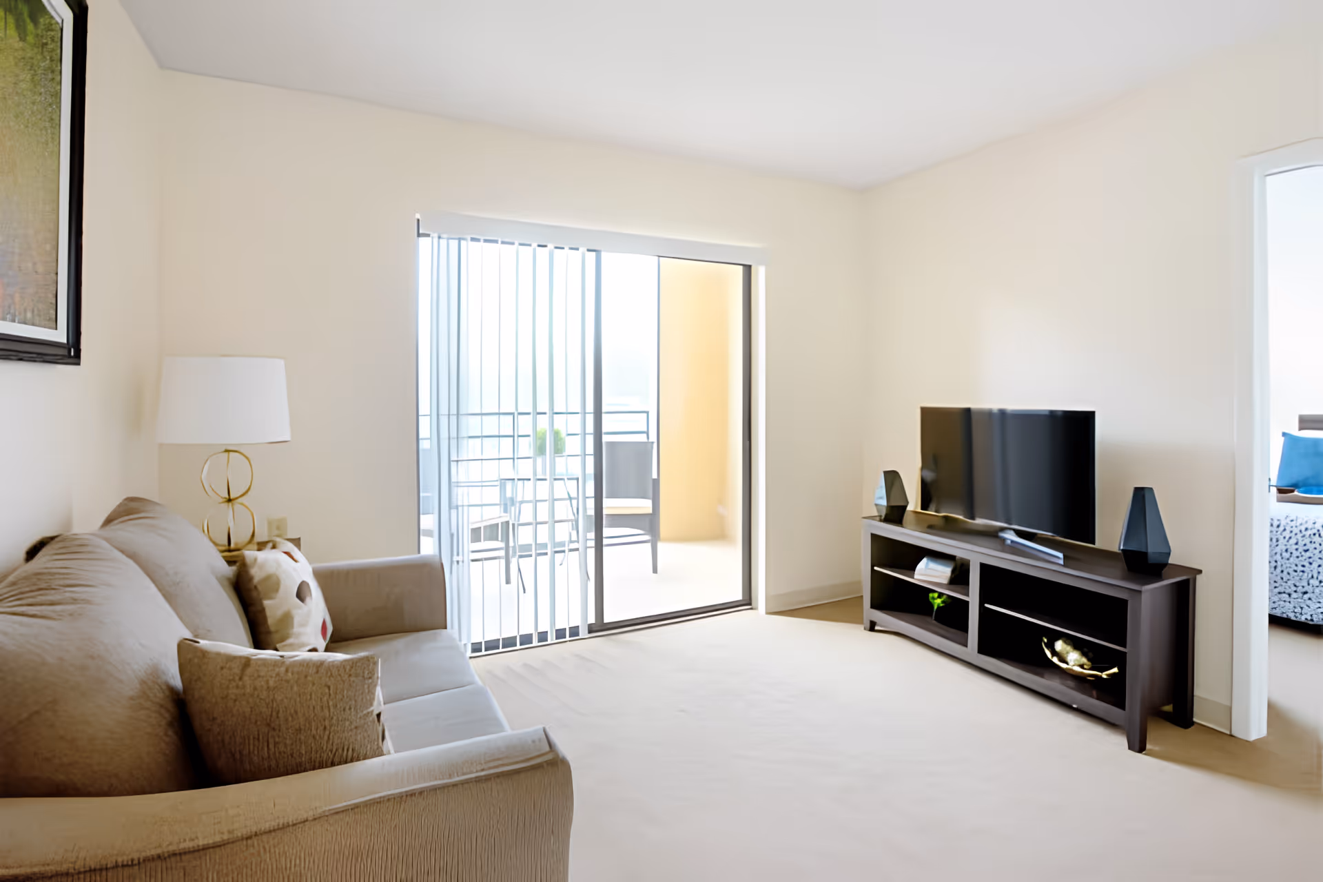 Bright living room with a beige sofa, TV on a dark stand, and sliding glass doors opening to a small balcony.