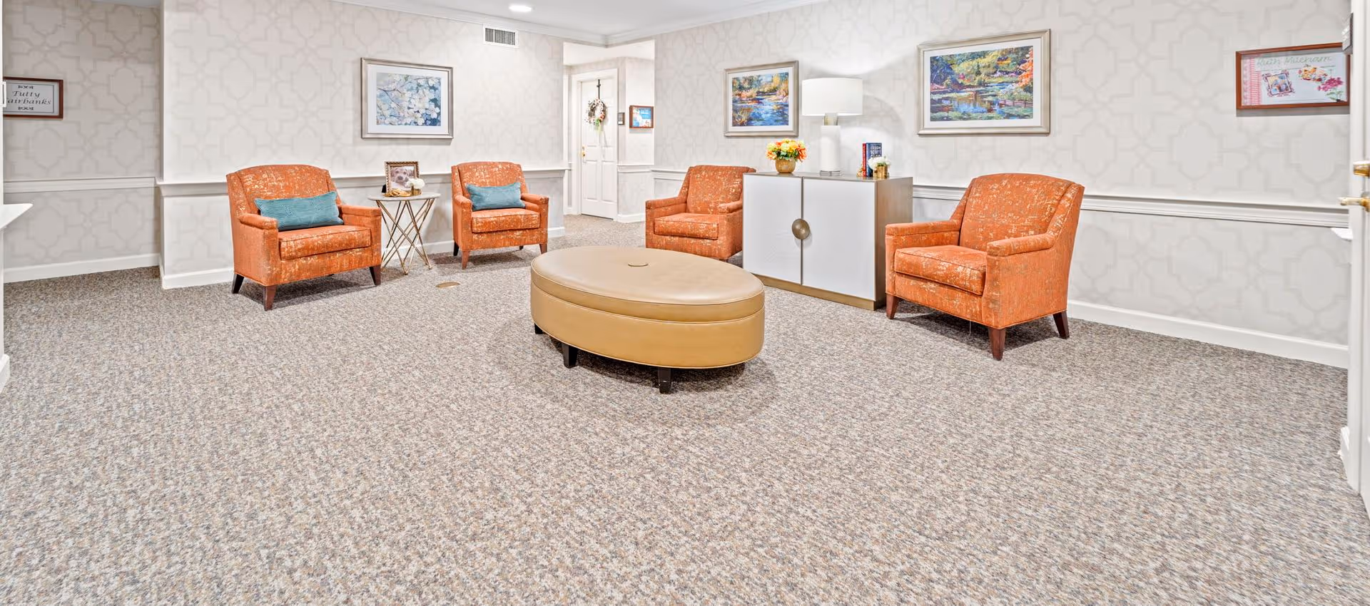 A cozy sitting area with four orange armchairs arranged around a large oval beige ottoman. The room has light patterned wallpaper, carpeted floor, and several framed paintings on the walls. A white cabinet with a lamp and decorative items is against one wall, and a door is visible in the background.