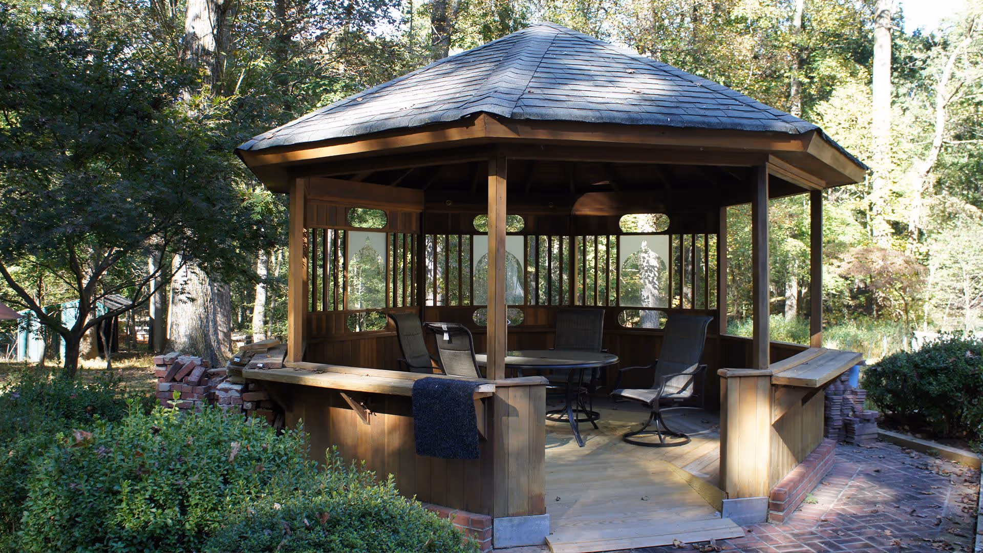 A wooden gazebo with a shingled roof situated in a garden area surrounded by trees and bushes. Inside the gazebo, there is a round table with four chairs. The setting is outdoors with natural sunlight filtering through the trees.