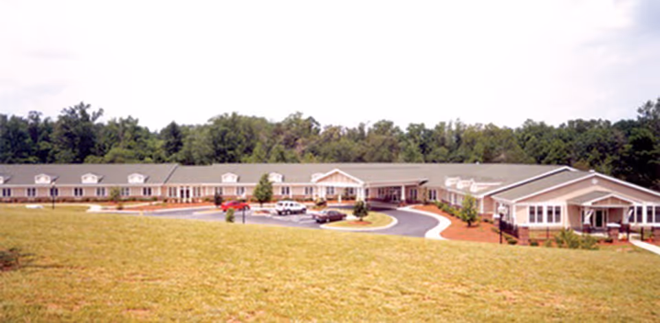 Long single-story senior living building with a circular driveway, grassy lawn in front and trees in the background.