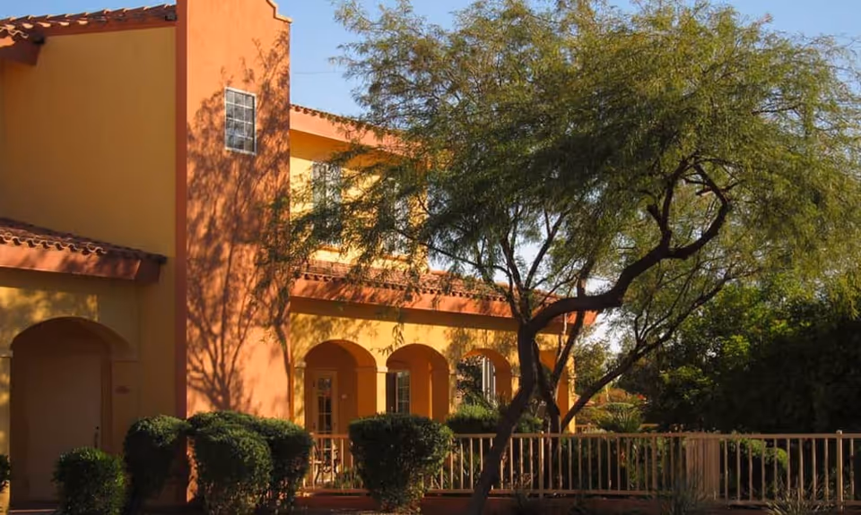 Exterior view of a two-story building with yellow and orange walls, arched doorways, and a tiled roof. The building is surrounded by green bushes and a tree with a curved trunk in the foreground under a clear blue sky.