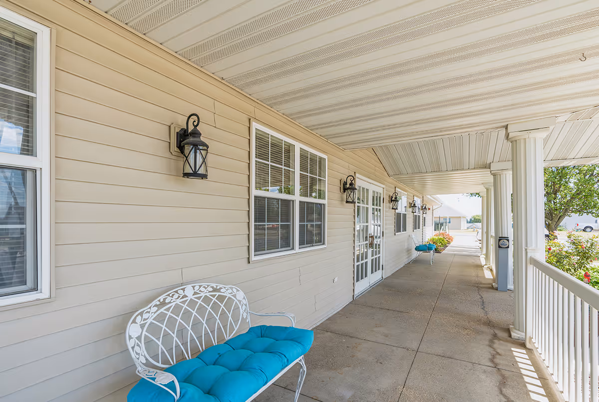 Covered outdoor walkway at a senior living facility with white railings and columns, beige siding walls, multiple windows, black lantern-style wall lights, and white metal benches with blue cushions.