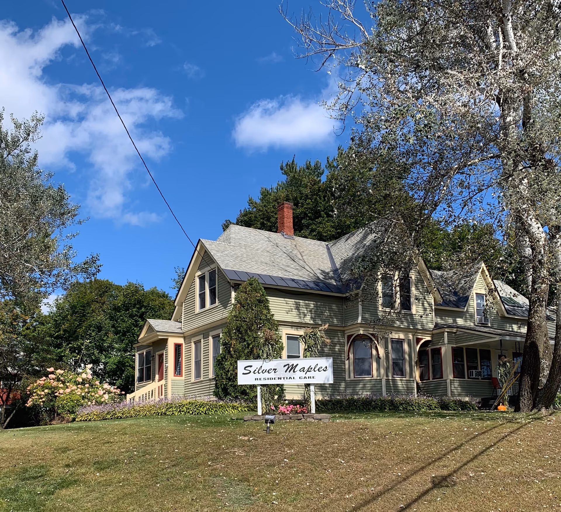 A large, light green residential house with a gray roof and red brick chimney under a blue sky with some clouds. There is a white sign in front of the house that reads 'Silver Maples Residential Care'. The house is surrounded by trees and a well-maintained lawn with some flowers near the sign.