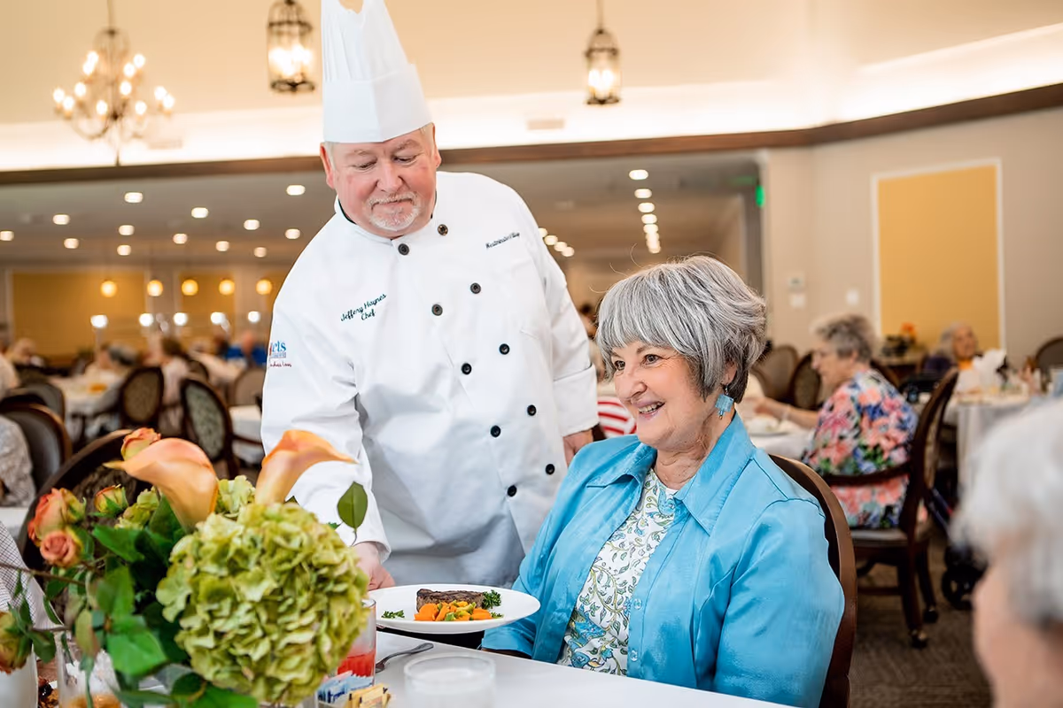 A chef in a white uniform and tall hat serves a plate of food to a smiling elderly woman seated at a dining table in a well-lit dining room with other seniors in the background.
