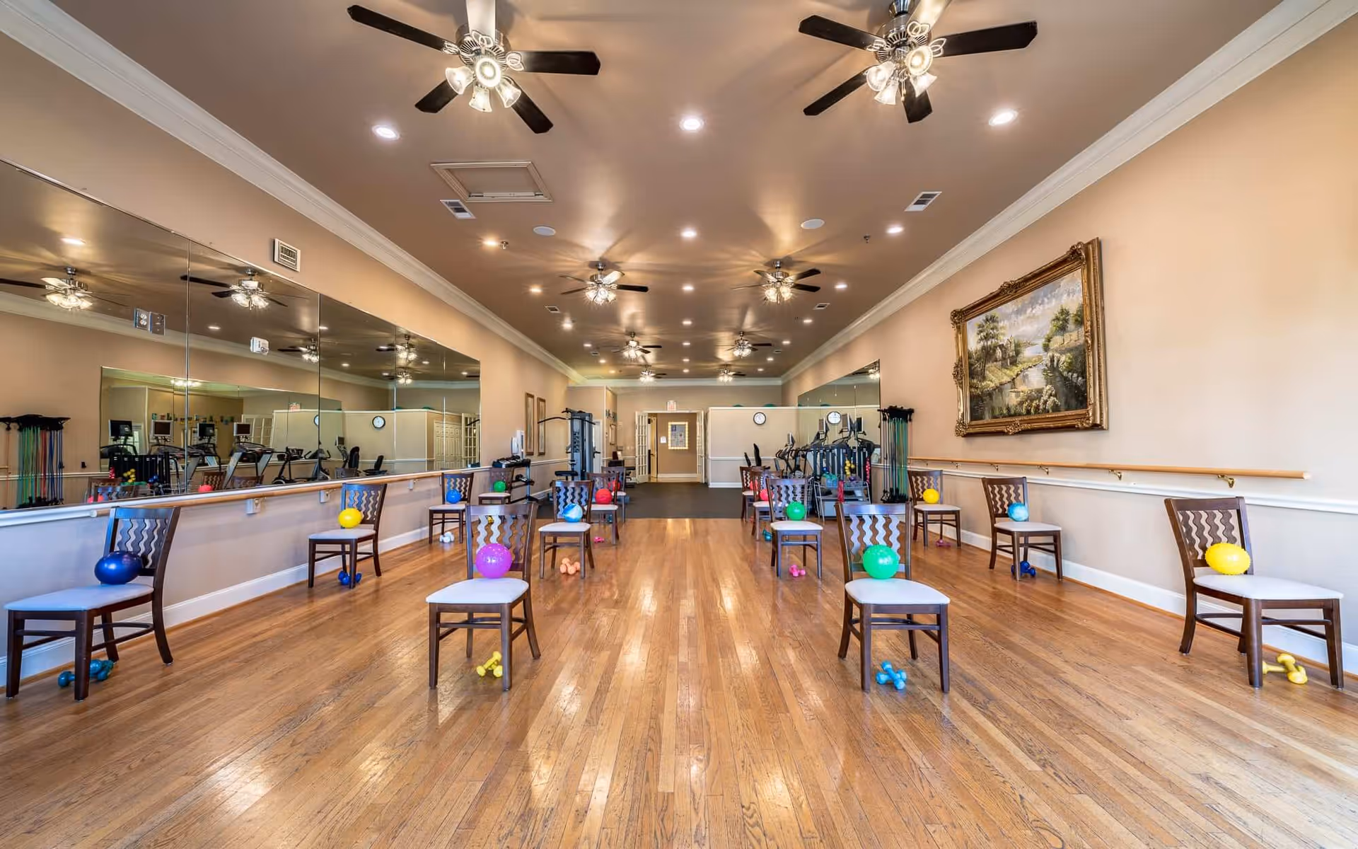A spacious exercise room with wooden floors, multiple chairs arranged in rows each holding a colorful exercise ball. Small dumbbells are placed on the floor near each chair. The room has large mirrors on one wall, ceiling fans with lights, and a large framed painting on the opposite wall. Exercise equipment is visible at the far end of the room.