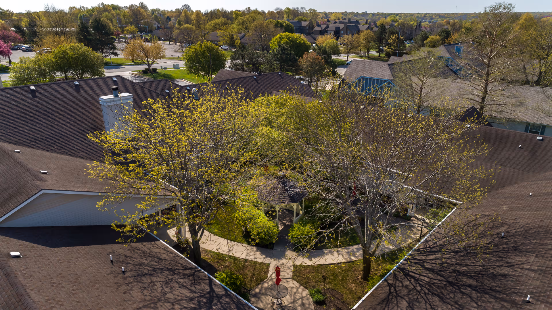 Aerial view of a senior living facility courtyard with a gazebo surrounded by trees and greenery. The courtyard is enclosed by the building's roof, and there are walking paths around the gazebo. In the background, there are more trees and residential houses.