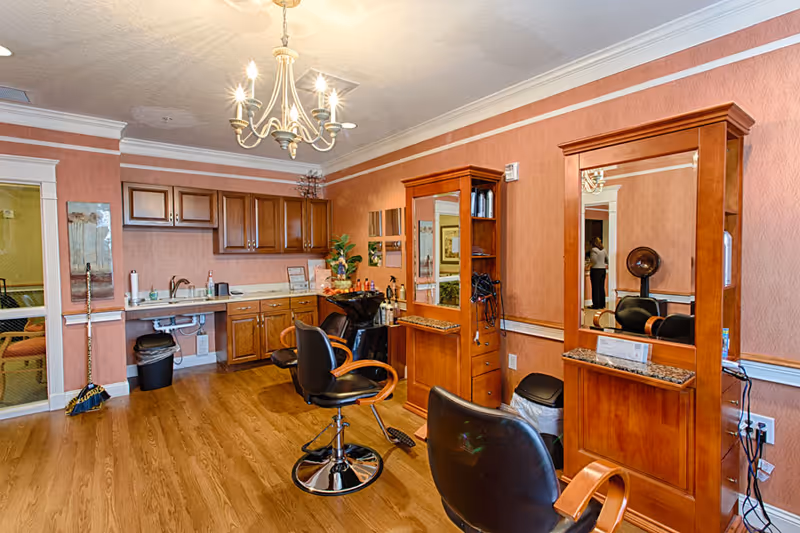 Interior view of a senior living facility's hair salon with wooden cabinetry, two styling stations with mirrors and chairs, a sink for washing hair, and a chandelier hanging from the ceiling. The room has wood flooring and peach-colored walls with white trim.