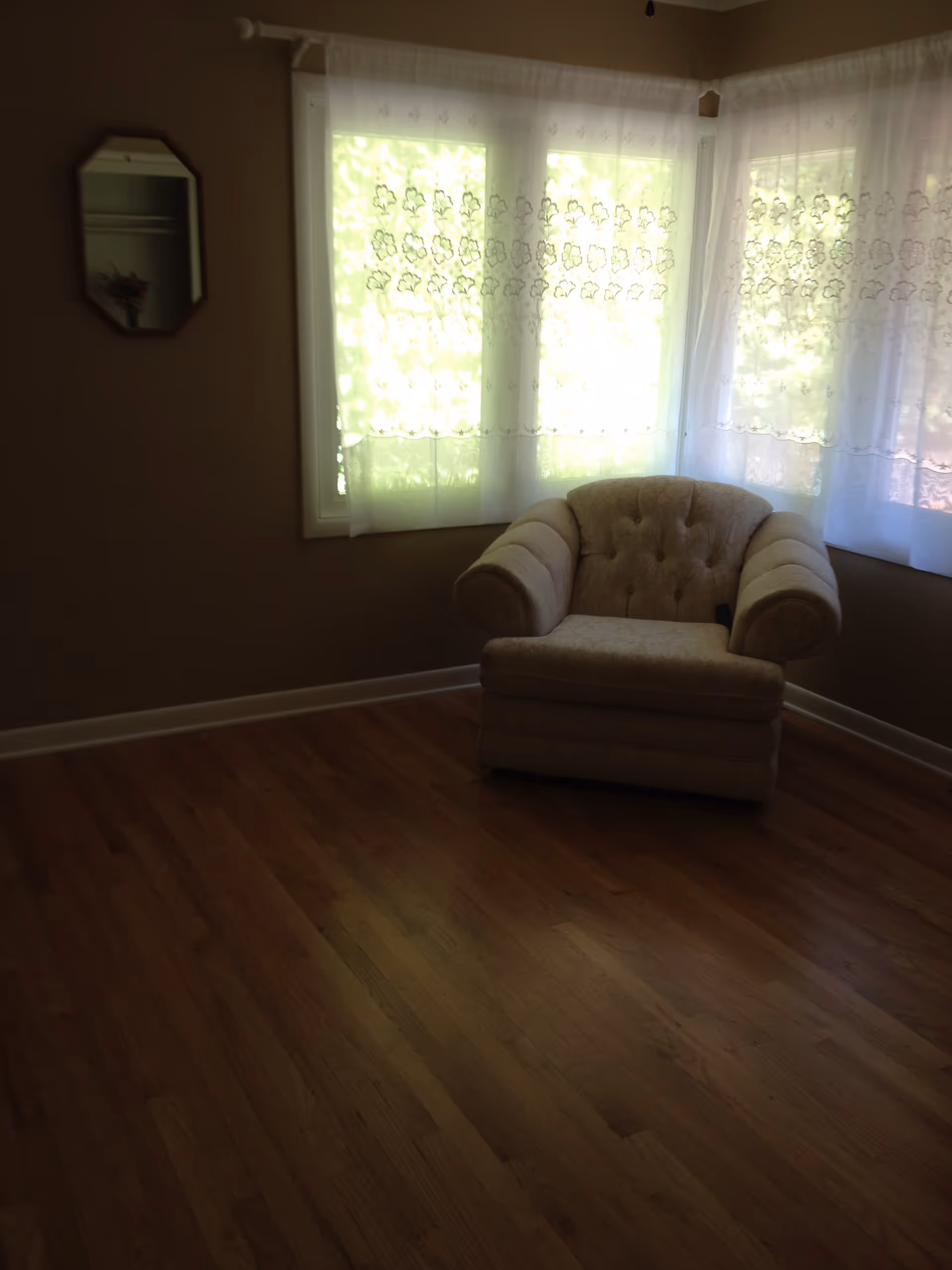 A cozy corner of a room with wooden flooring, featuring a single beige upholstered armchair placed near two windows covered with sheer white curtains with floral patterns. A small mirror hangs on the adjacent wall.