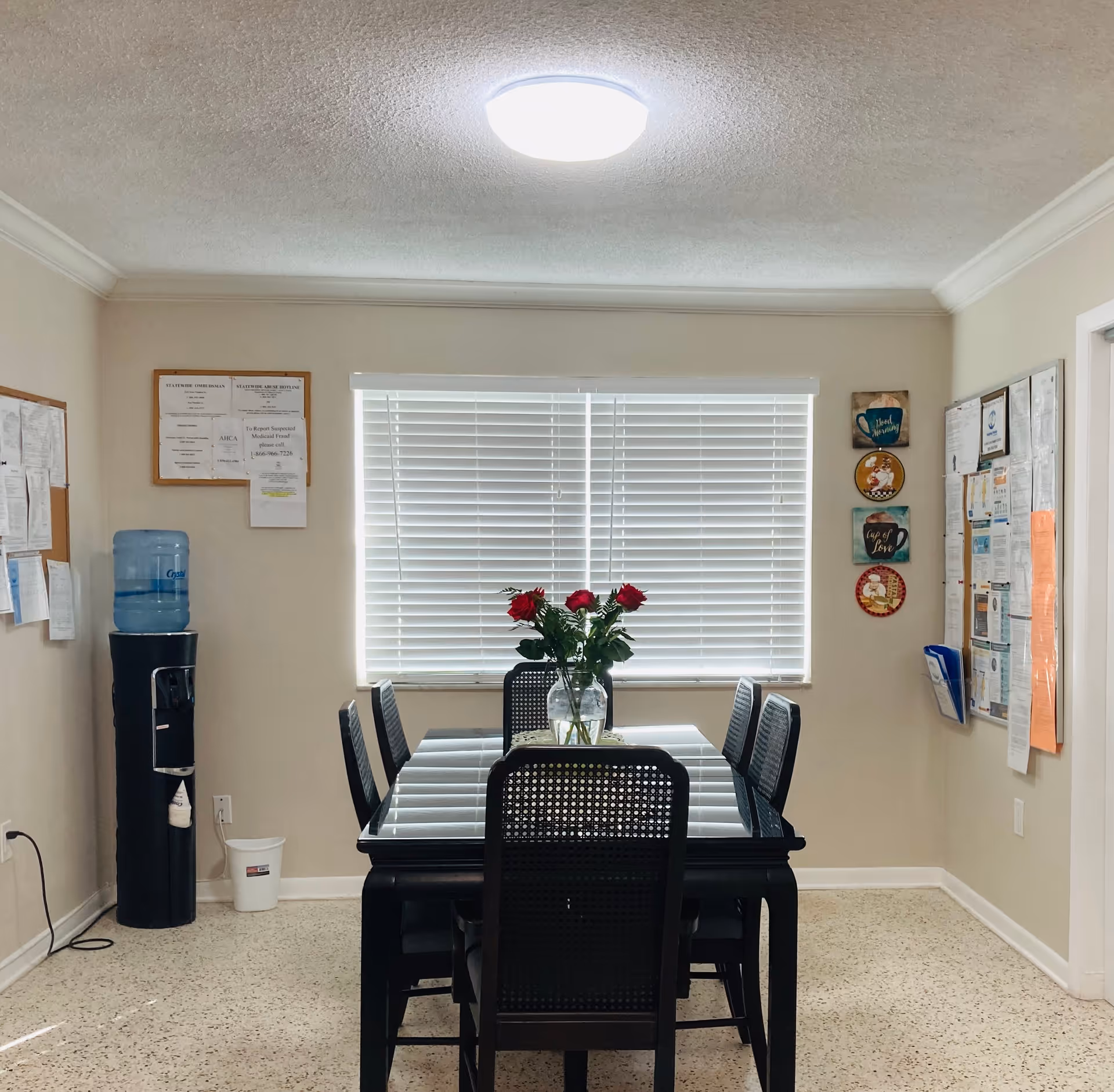 A small dining area with a black glass-top table and six black chairs. A vase with red roses is placed in the center of the table. The room has beige walls with two bulletin boards filled with papers on either side of a window with white blinds. A water cooler is positioned to the left of the window.