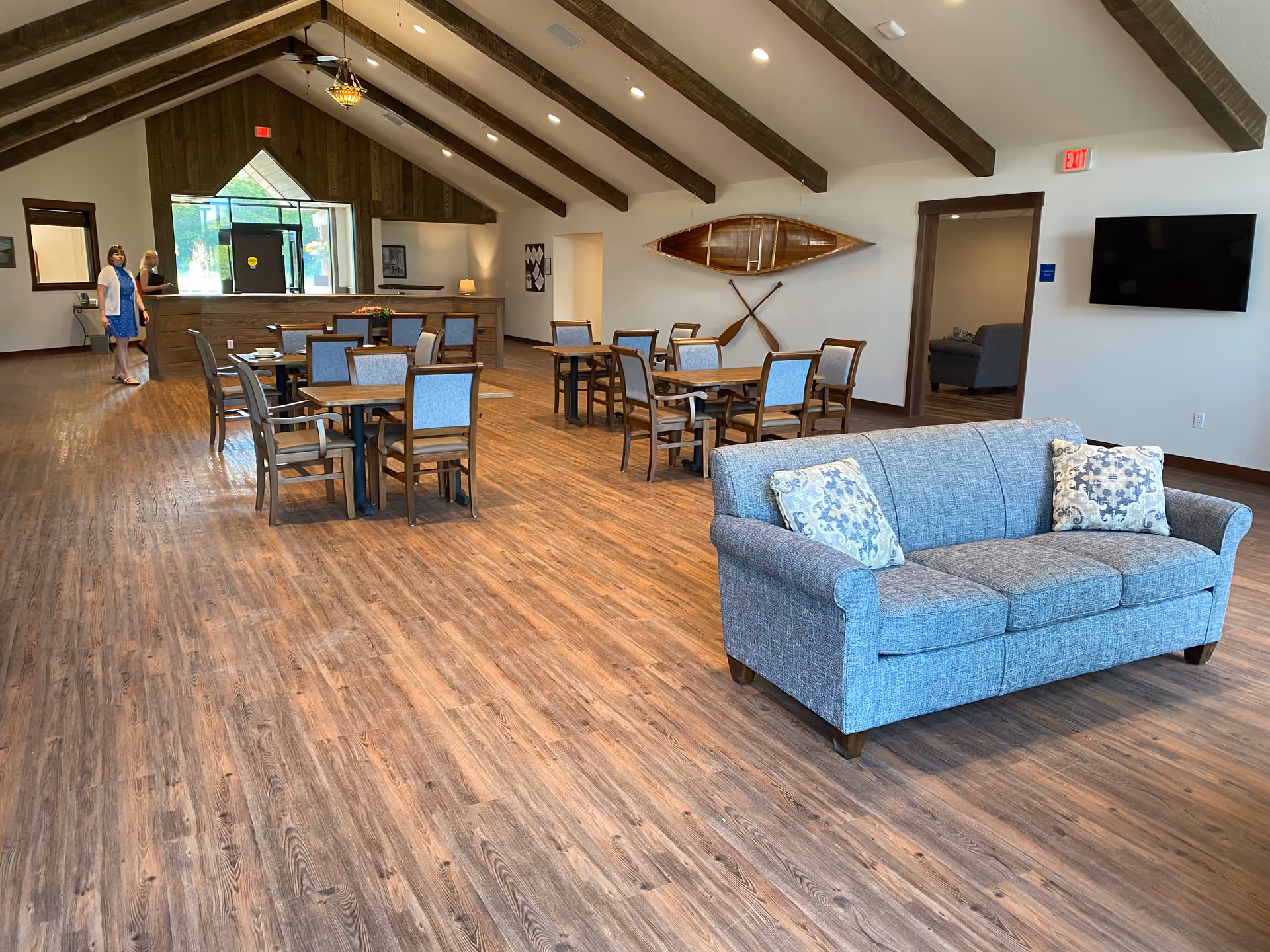 A spacious senior living common area with wooden floors and exposed ceiling beams. The room features several tables with chairs arranged for dining or socializing, a gray upholstered sofa with patterned pillows in the foreground, a wall-mounted TV, and a decorative wooden canoe and paddles mounted on the wall. Two women are standing near the entrance area in the background.