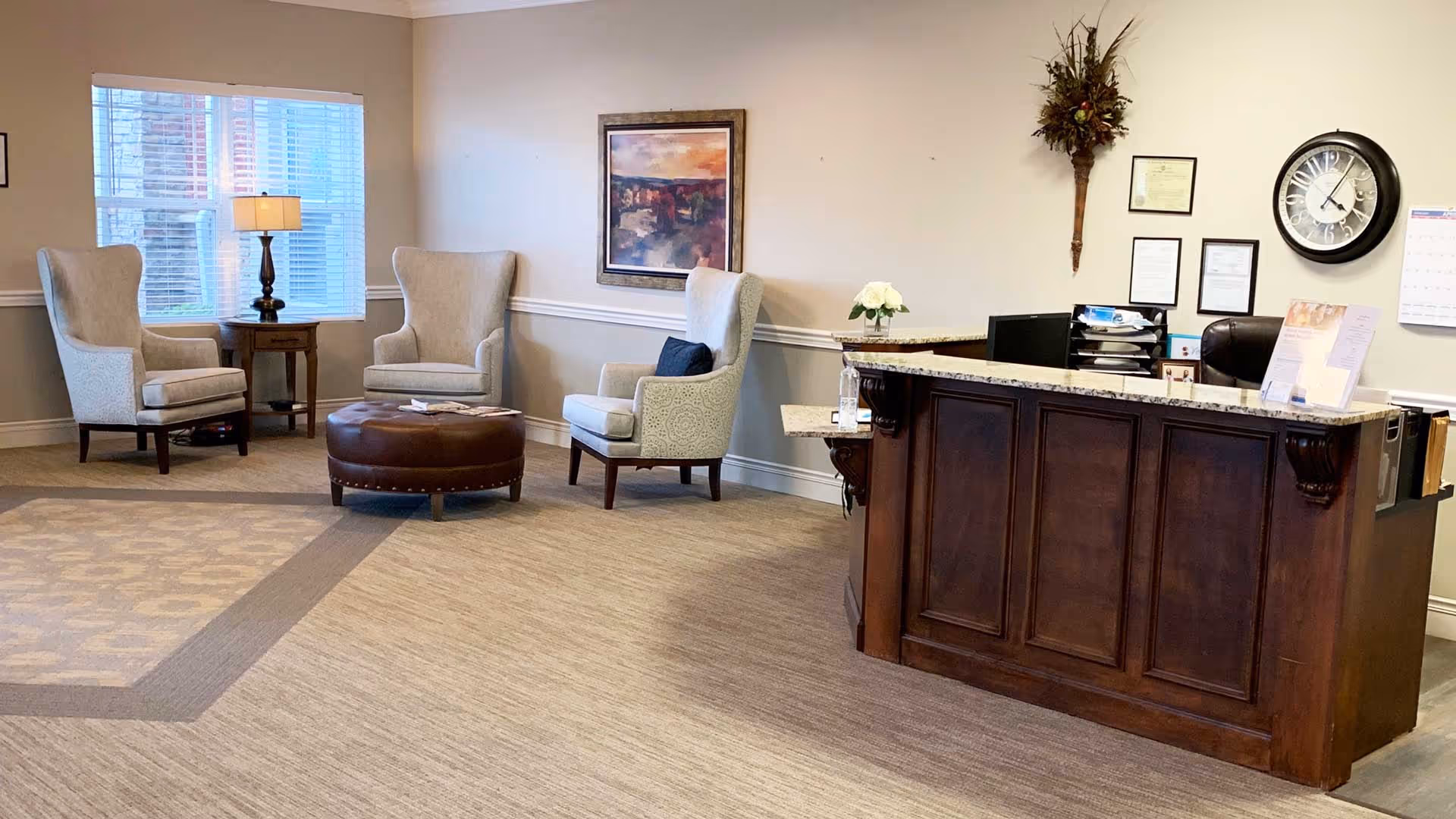 Reception area of Maristone of Providence featuring a wooden front desk with a granite countertop, a clock and framed documents on the wall behind it, and a seating area with three upholstered armchairs, a round leather ottoman, a side table with a lamp, and a window with blinds.