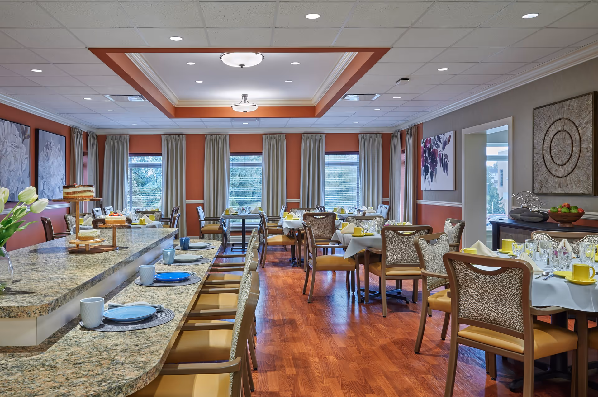 A well-lit dining room in a senior living facility with multiple tables set with yellow cups, plates, and napkins. There is a long granite countertop with chairs and cakes displayed on a tiered stand. The room features large windows with beige curtains, wooden flooring, and decorative wall art.