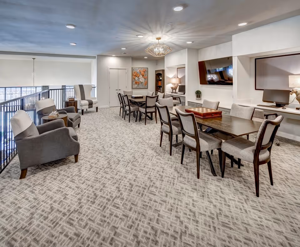 A spacious, well-lit common area in a senior living facility featuring multiple seating arrangements including armchairs and dining chairs around tables. The room has a patterned carpet, a modern chandelier, a wall-mounted TV, and computer workstations built into the wall. There is artwork on the wall and a railing overlooking a lower level.