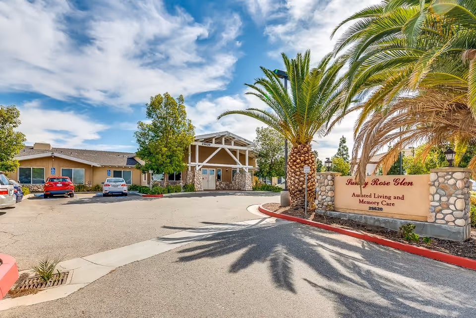 Exterior view of Sunny Rose Glen Assisted Living and Memory Care facility with a parking lot, palm trees, and a clear blue sky with some clouds.