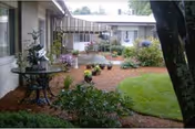 Small courtyard with a lawn, flowerbeds, potted plants and a round metal table between single-story building wings.