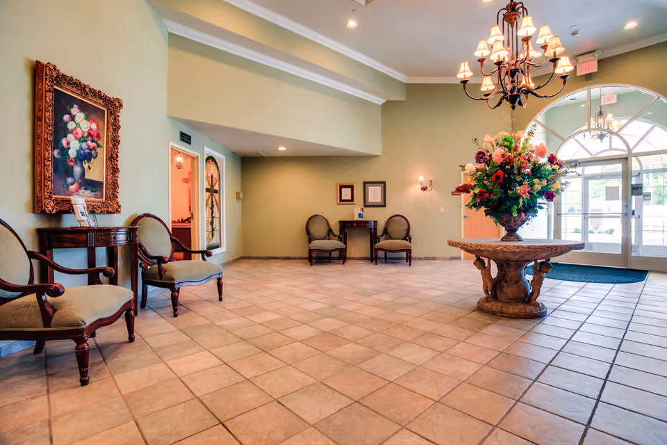 Spacious and well-lit lobby area with tiled floor, several upholstered chairs along the walls, a large ornate table in the center holding a vibrant floral arrangement, a chandelier hanging from the ceiling, framed artwork on the walls, and glass double doors leading outside.