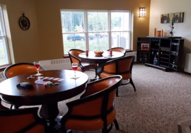 Carpeted communal dining room with round wooden tables, orange-upholstered chairs, wine glasses on the tables, and a cabinet under wall art by a large window.