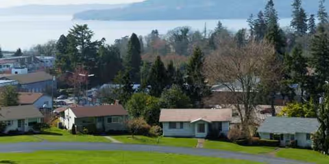 Suburban neighborhood with front lawns, trees, and small single-story houses along a curved road.