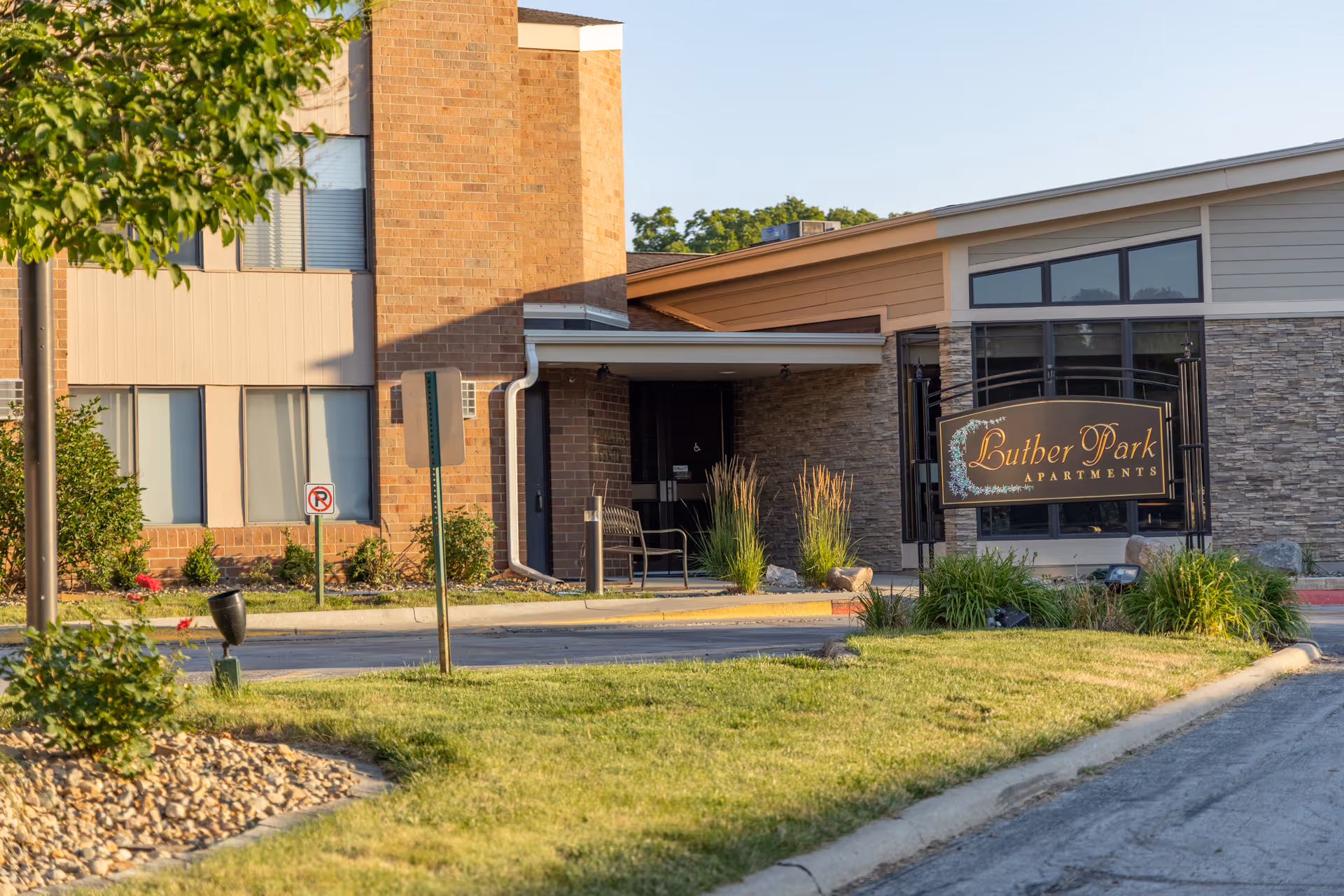 Exterior view of Luther Park Apartments building entrance with a sign displaying the community name, surrounded by grass, plants, and a tree.