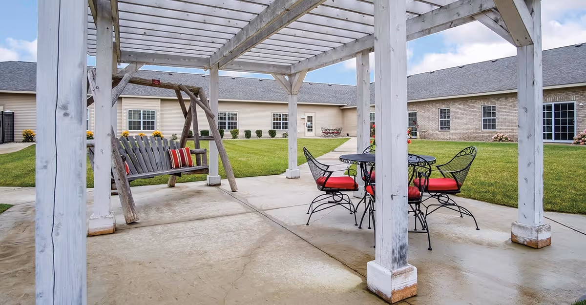 Outdoor patio area at Heritage Hill Assisted Living & Memory Care featuring a white wooden pergola, a wooden swing with red and white striped cushions, and a black metal table with four chairs that have red cushions. The patio is surrounded by a well-maintained lawn and buildings with beige siding and brick walls under a partly cloudy sky.
