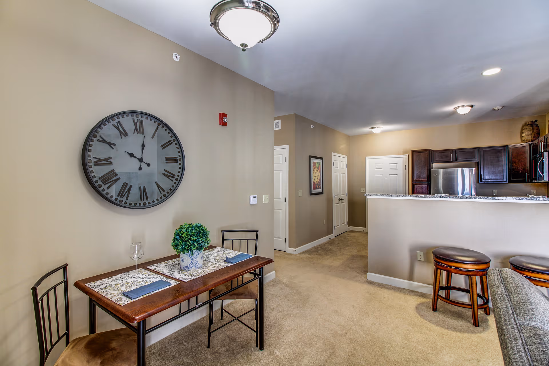 Interior view of a senior living facility showing a small dining area with a wooden table set for two, a large wall clock with Roman numerals, and a kitchen with dark wood cabinets and stainless steel appliances in the background. The space has beige walls and carpeted floors, with bar stools at the kitchen counter.