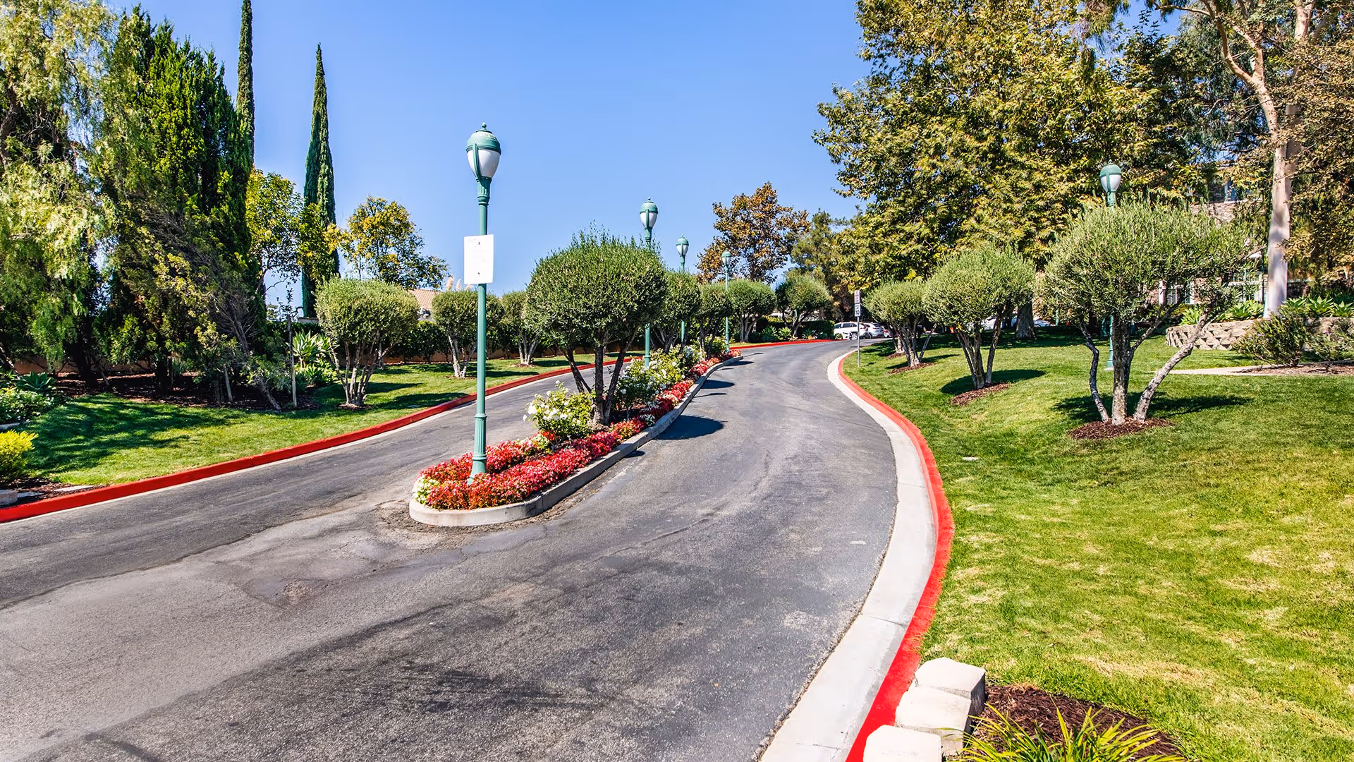A paved driveway lined with red curbs curves through a landscaped area with green grass, trimmed bushes, and tall trees under a clear blue sky. Several green street lamps are positioned along the driveway.