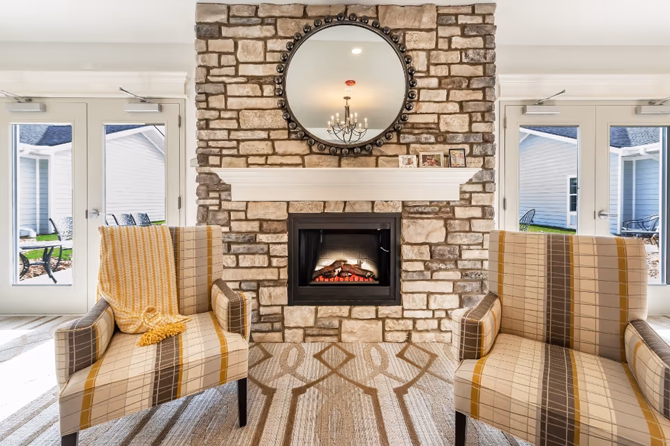 Two plaid armchairs face a stone fireplace with a round mirror above, flanked by glass doors to the outside.