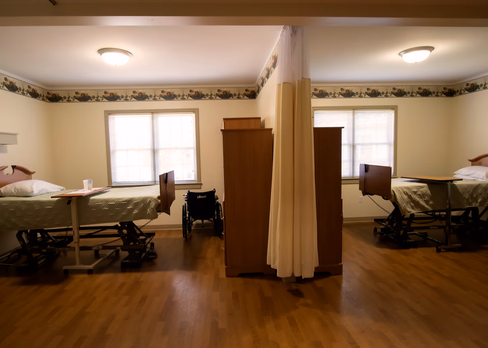 A shared room in a senior living facility with two hospital-style beds separated by a wooden divider and a privacy curtain. Each bed has a green quilted blanket and an overbed table. A wheelchair is positioned near the divider. The room has wooden flooring, two windows with blinds, and a floral border near the ceiling.