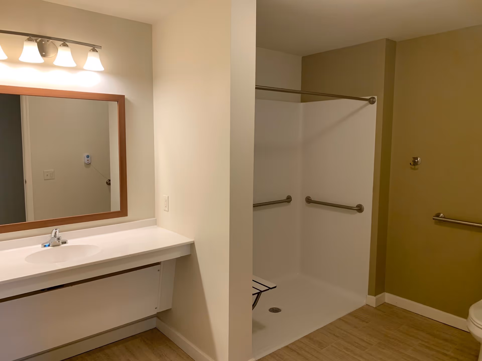 A bathroom with a white sink and countertop on the left side, a large mirror with wooden frame above the sink, and a light fixture with four lights above the mirror. On the right side, there is a walk-in shower with grab bars and a foldable shower seat. The floor has light wood-like tiles, and the walls are painted in neutral tones.