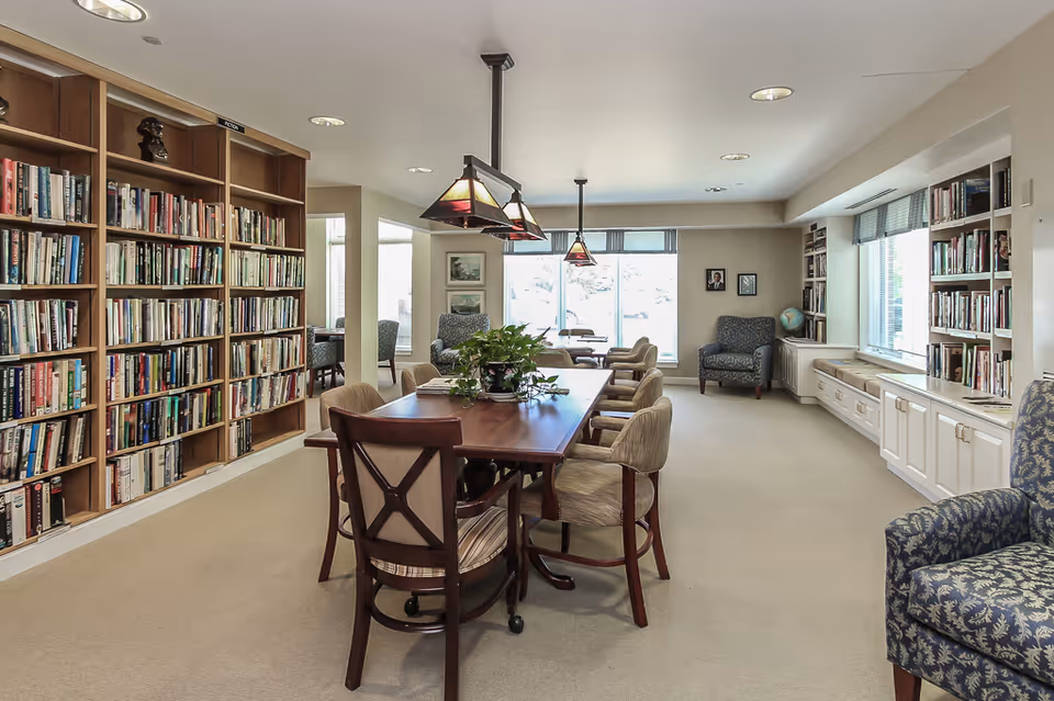 A bright and spacious library room with large bookshelves filled with books along the left and right walls. In the center, there is a wooden table surrounded by several cushioned chairs. Two pendant lights hang above the table. There are additional armchairs near the windows at the far end of the room, which allow natural light to fill the space.