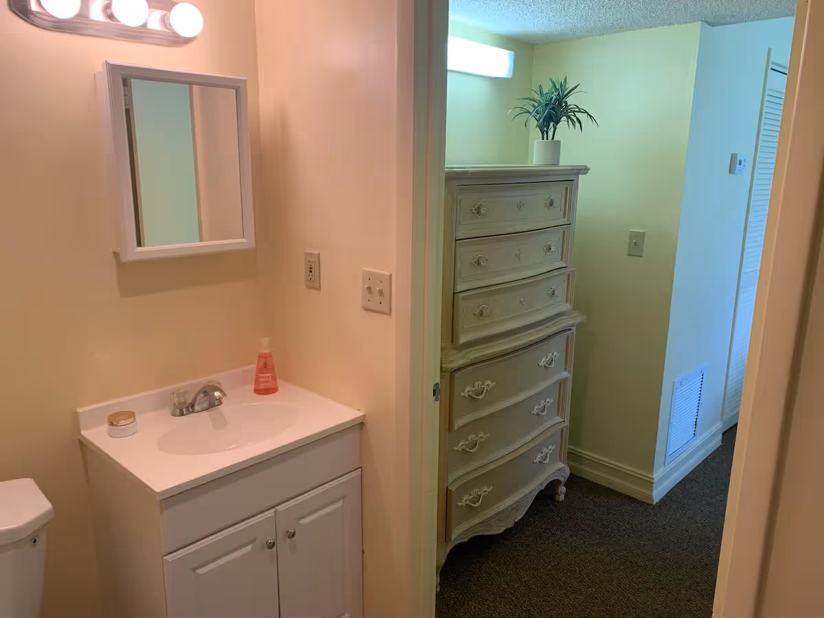 Small bathroom vanity with sink and mirror next to an open doorway revealing a hallway and a tall dresser.
