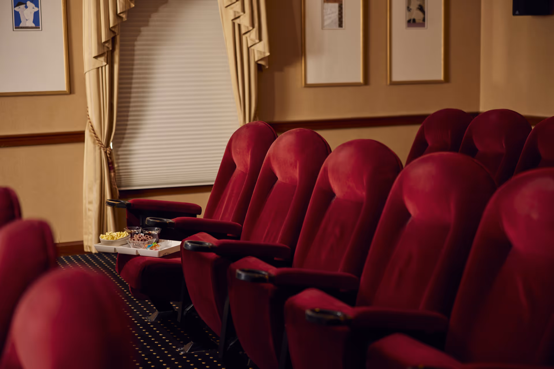 A row of plush red theater-style seats in a cozy room with beige walls and framed artwork. A tray with popcorn, candy, and snacks is placed on one of the armrests. The window is covered with a closed blind and beige curtains.