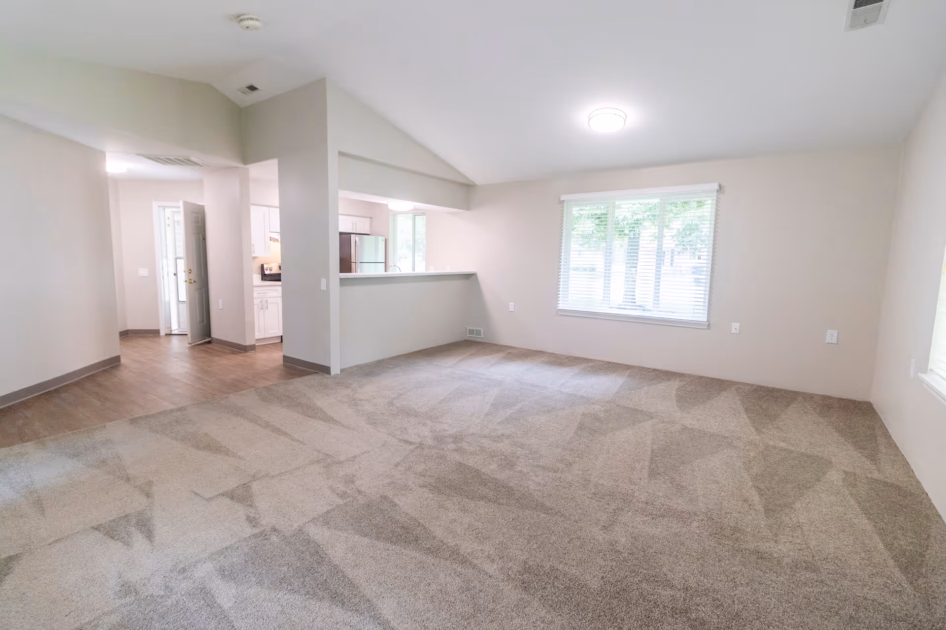 An empty living room area with beige carpet and light gray walls. There is a large window with white blinds letting in natural light. The room opens into a kitchen area with white cabinets and stainless steel appliances. The flooring transitions from carpet in the living room to wood or laminate in the kitchen and entryway. The ceiling has a modern light fixture and a vent.