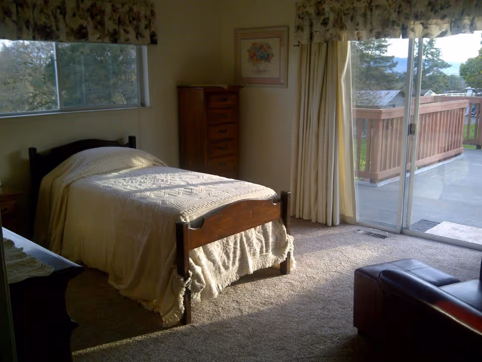 A bedroom with a single wooden bed covered with a cream-colored textured blanket. There is a wooden dresser with drawers against the wall and a framed floral picture above it. The room has carpeted flooring and large windows with floral valances and curtains. A sliding glass door opens to a wooden balcony with a view of trees and distant hills. A dark leather ottoman is partially visible in the foreground.