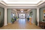 Spacious lobby area with tiled floor, green walls, white columns, and a chandelier. There are plants and decorative items along the walls, and glass doors at the far end leading outside.