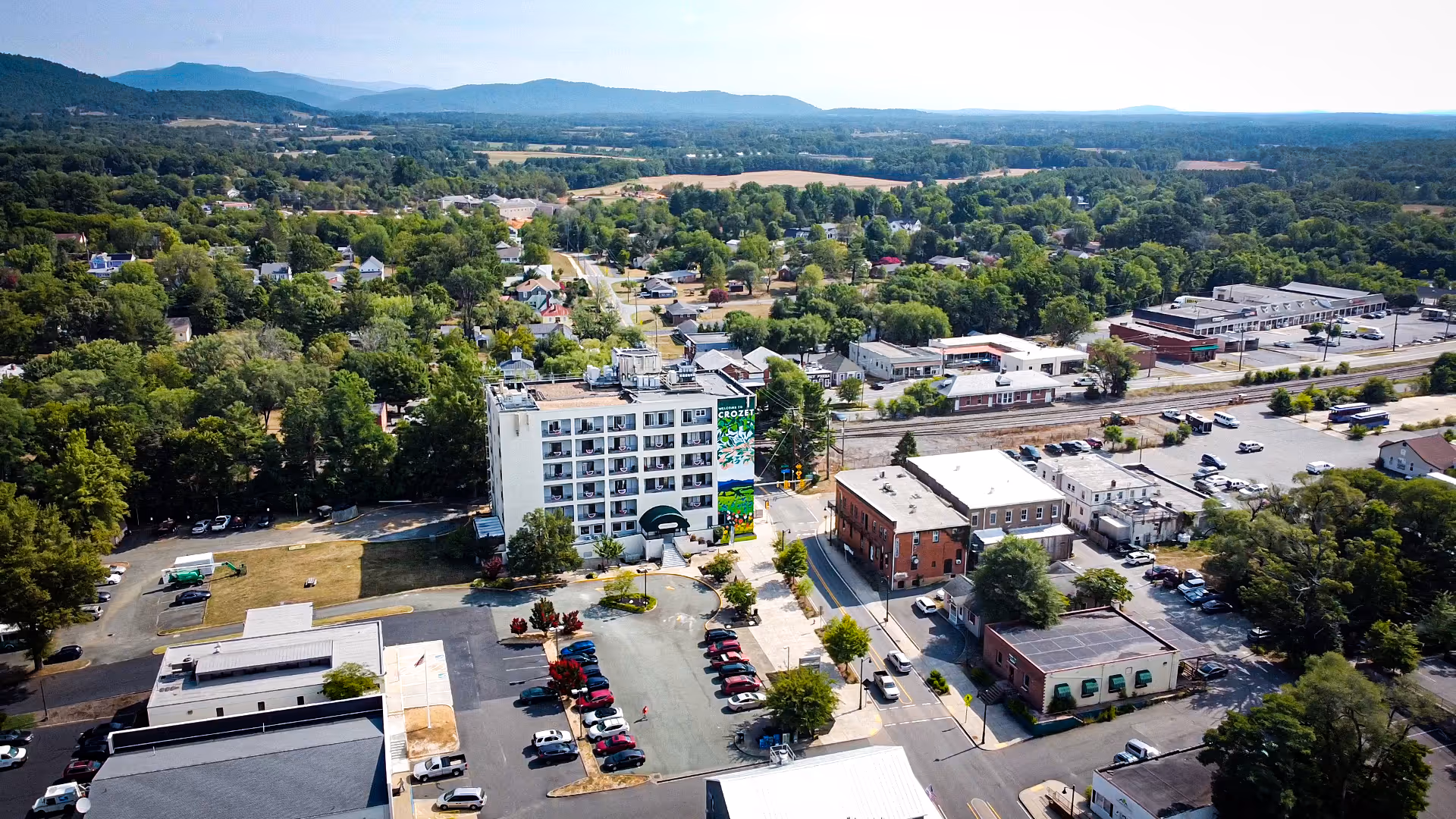 Aerial view of the English Meadows Crozet Campus showing a multi-story senior living building, parking lot, nearby commercial buildings and tree-lined residential areas with hills in the background.