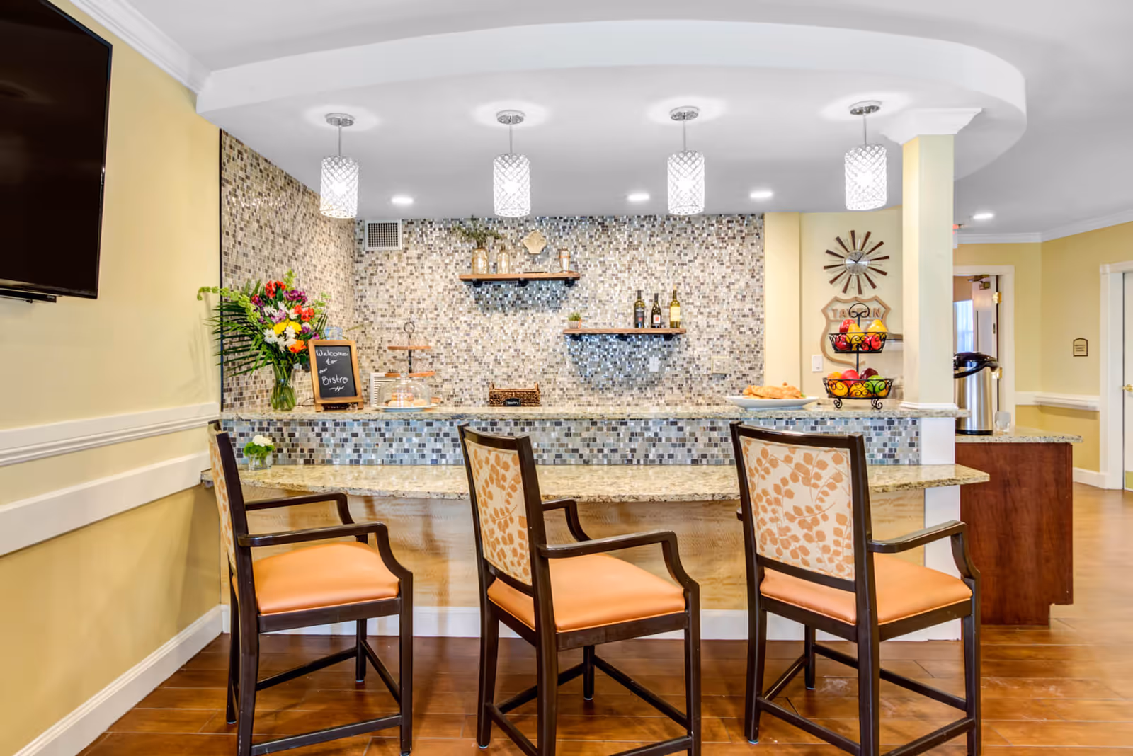A cozy bistro area in a senior living facility with a granite countertop bar and three high chairs with orange cushions and patterned backs. The wall behind the bar features a mosaic tile backsplash with two wooden shelves holding bottles and decorative items. Four pendant lights hang from the ceiling above the bar. A vase with colorful flowers and a small chalkboard sign that says 'Welcome to Bistro' are on the left side of the counter. On the right side, there is a tiered fruit basket and a plate of croissants. The room has warm yellow walls and wood flooring.