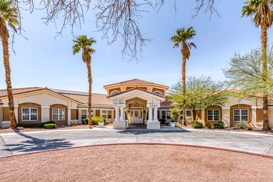 Front exterior view of a single-story building with a covered entrance supported by columns, surrounded by palm trees and desert landscaping under a clear blue sky.