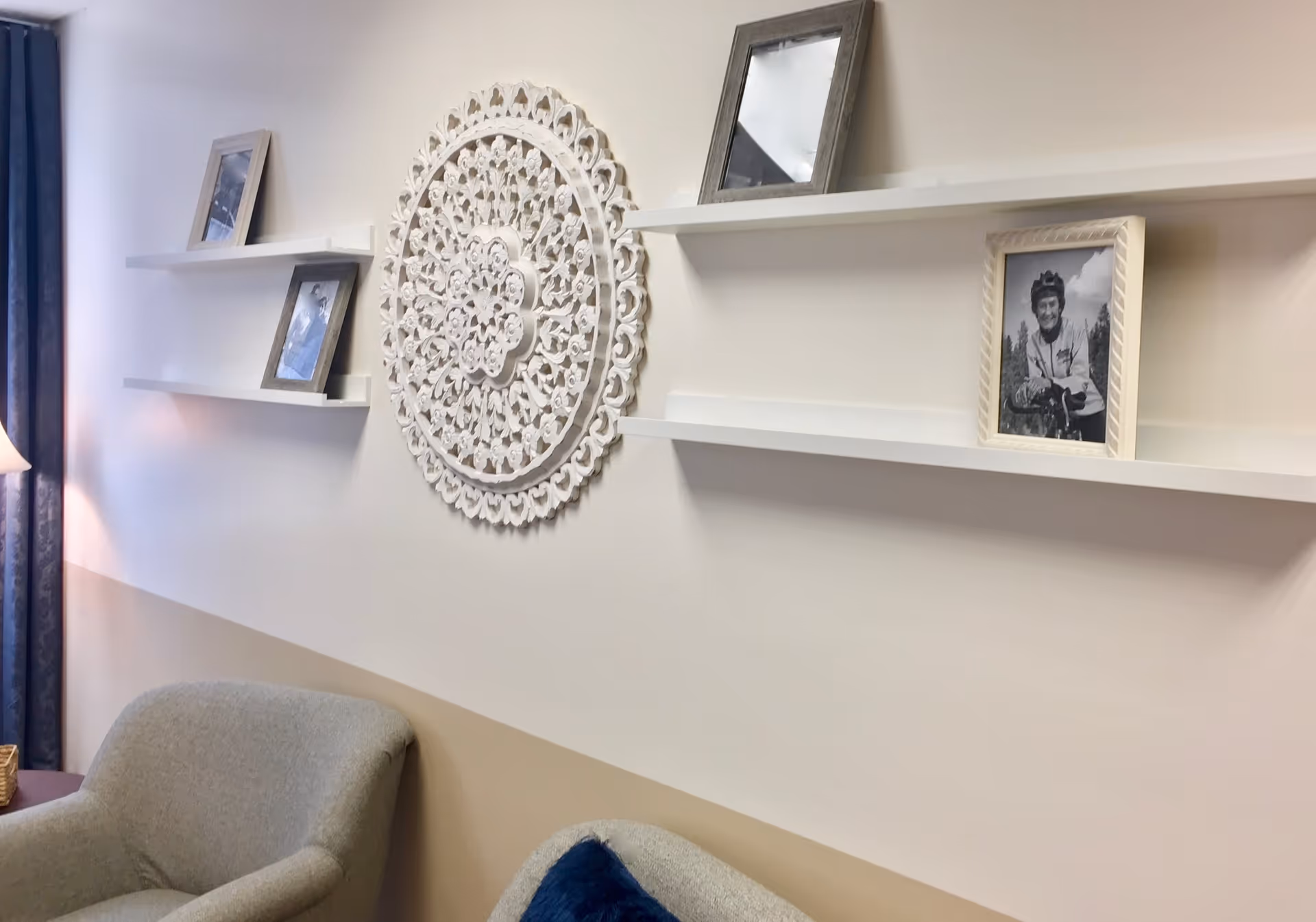 Two upholstered chairs beneath white floating shelves holding framed photos and a decorative round wall medallion.
